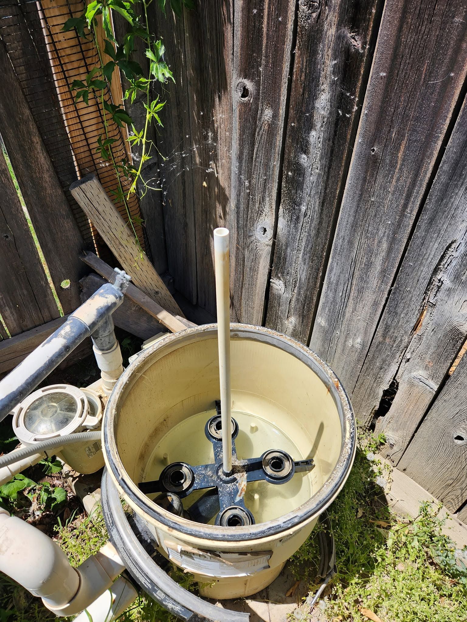 White plastic utility bucket with pipes and wires beside a weathered wooden fence outdoors