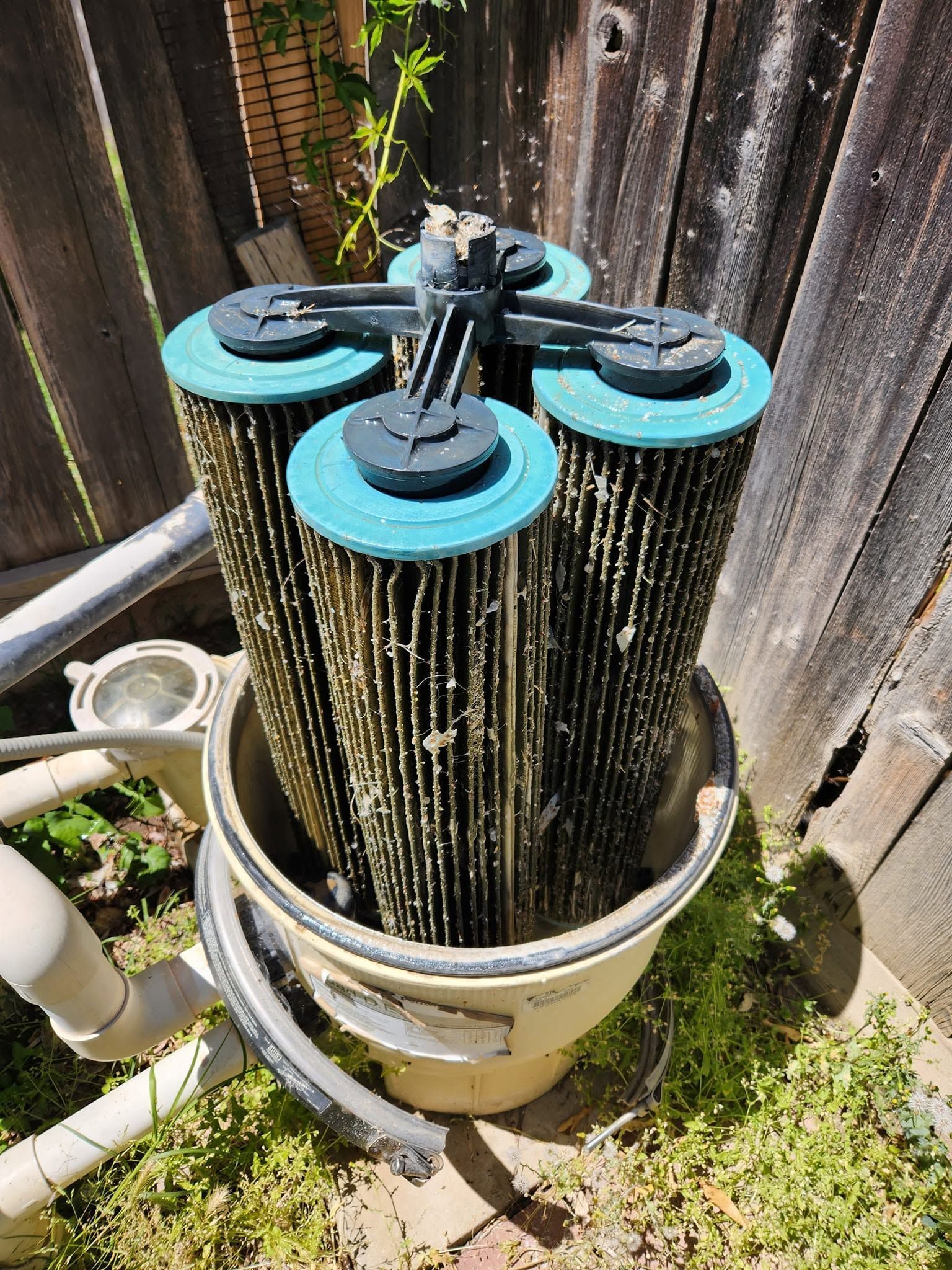 Cylindrical pool filter cartridge with blue caps installed in a white housing outdoors beside a wooden fence