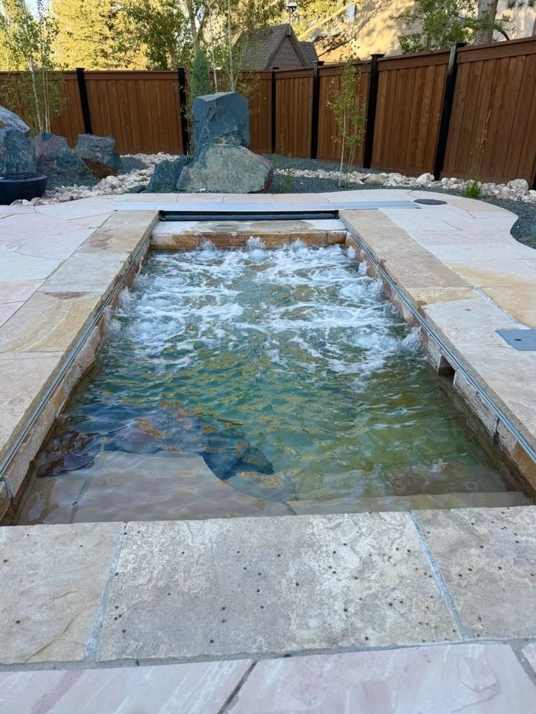 Backyard stone water feature with a shallow pool and bubbling fountain, bordered by a wooden fence.