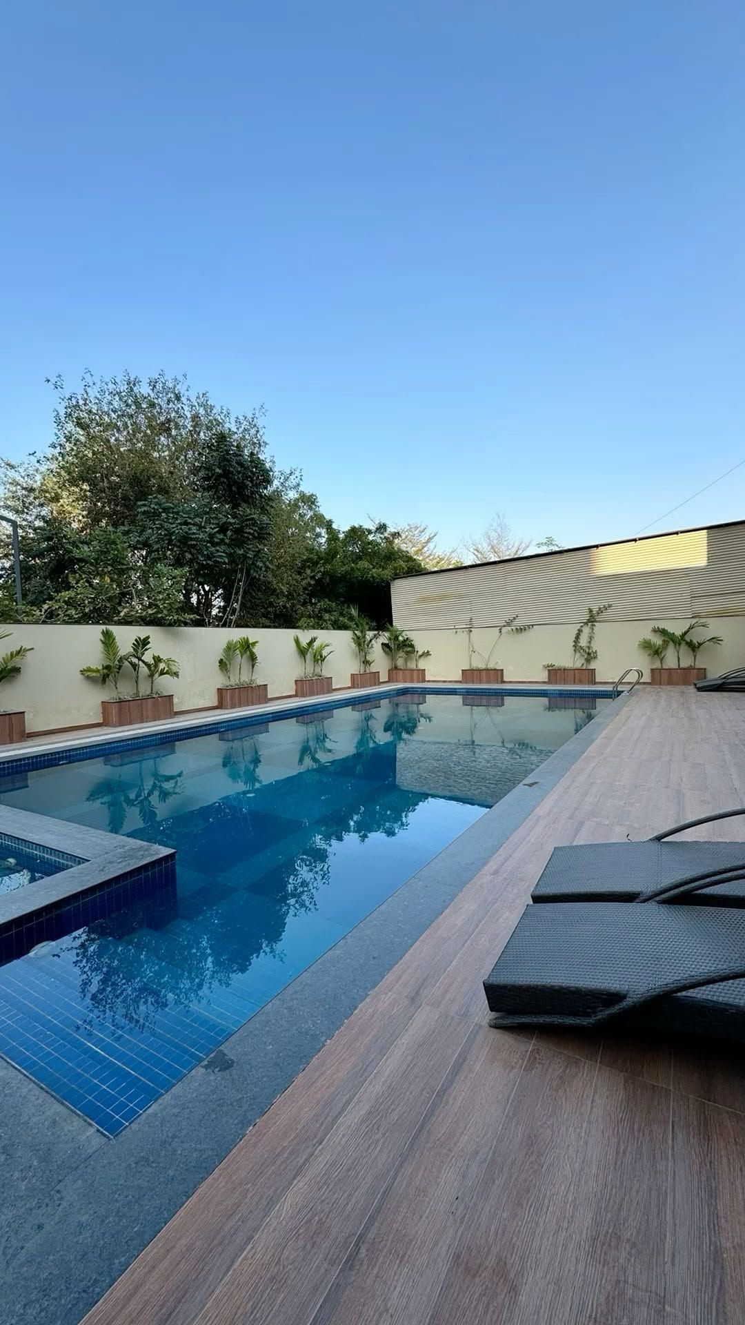 Backyard swimming pool with blue water, wooden deck, lounge chairs, and potted plants under a clear sky