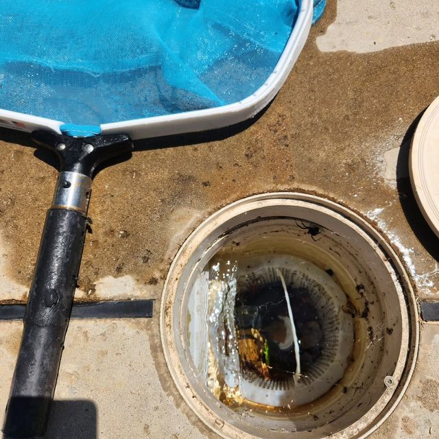 Blue pool skimmer net beside an open circular drain in concrete pavement