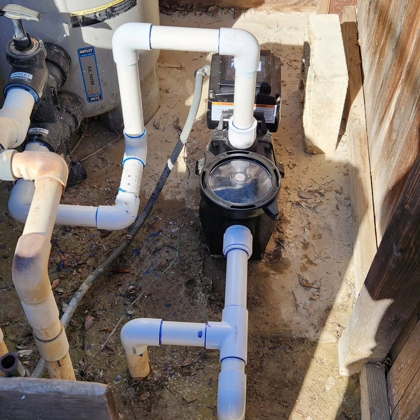Outdoor pool plumbing with white PVC pipes and a black pump beside a wall and dirt ground