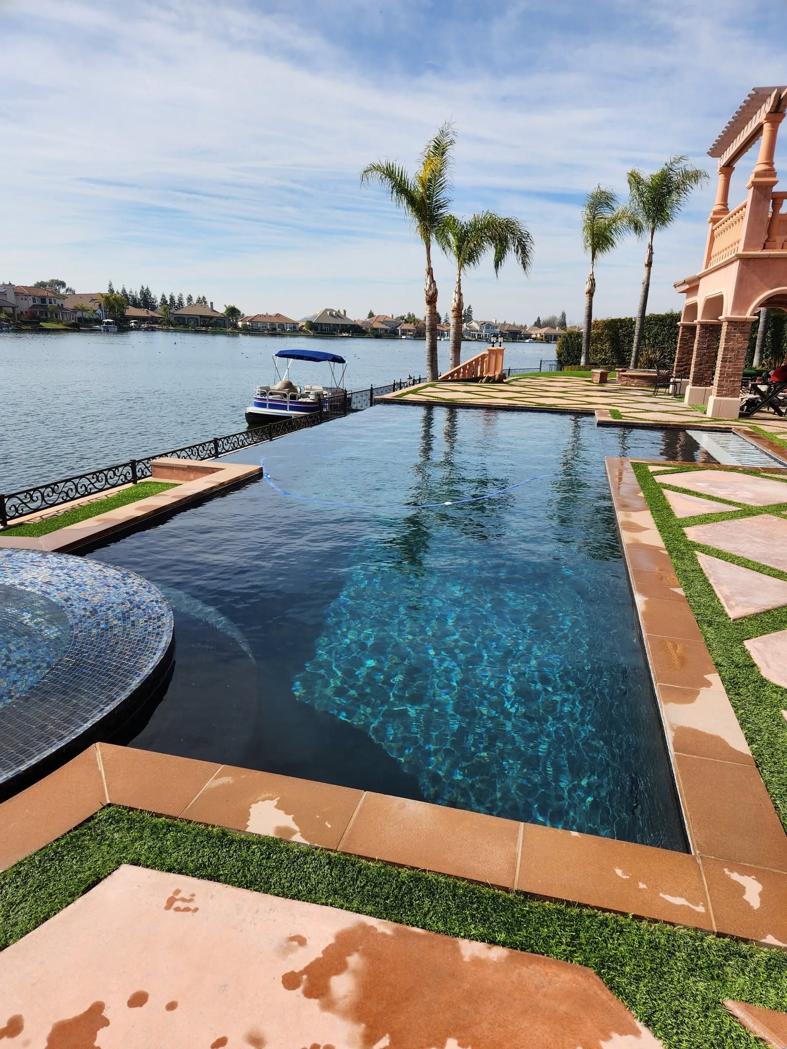 Luxury infinity pool beside a waterfront patio with palm trees and boats on a sunny day