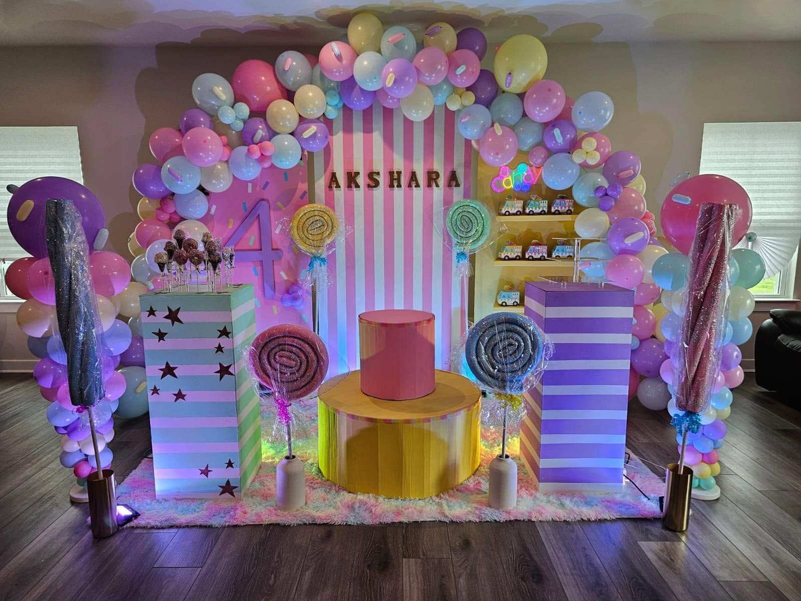 Colorful balloon-decorated stage with “AKSHARA” sign, pink and purple accents, and pedestal tables