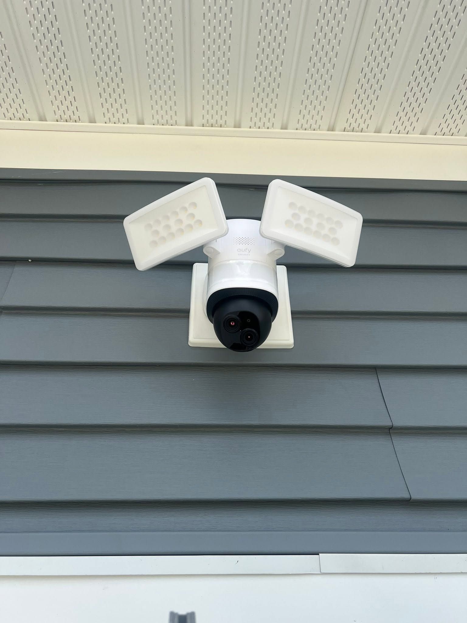 Outdoor security camera with two motion lights mounted on gray siding under a roof overhang