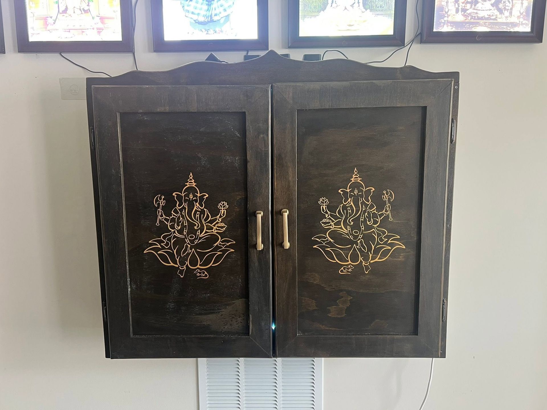 Black wall cabinet with gold floral designs and two doors hanging beneath framed pictures
