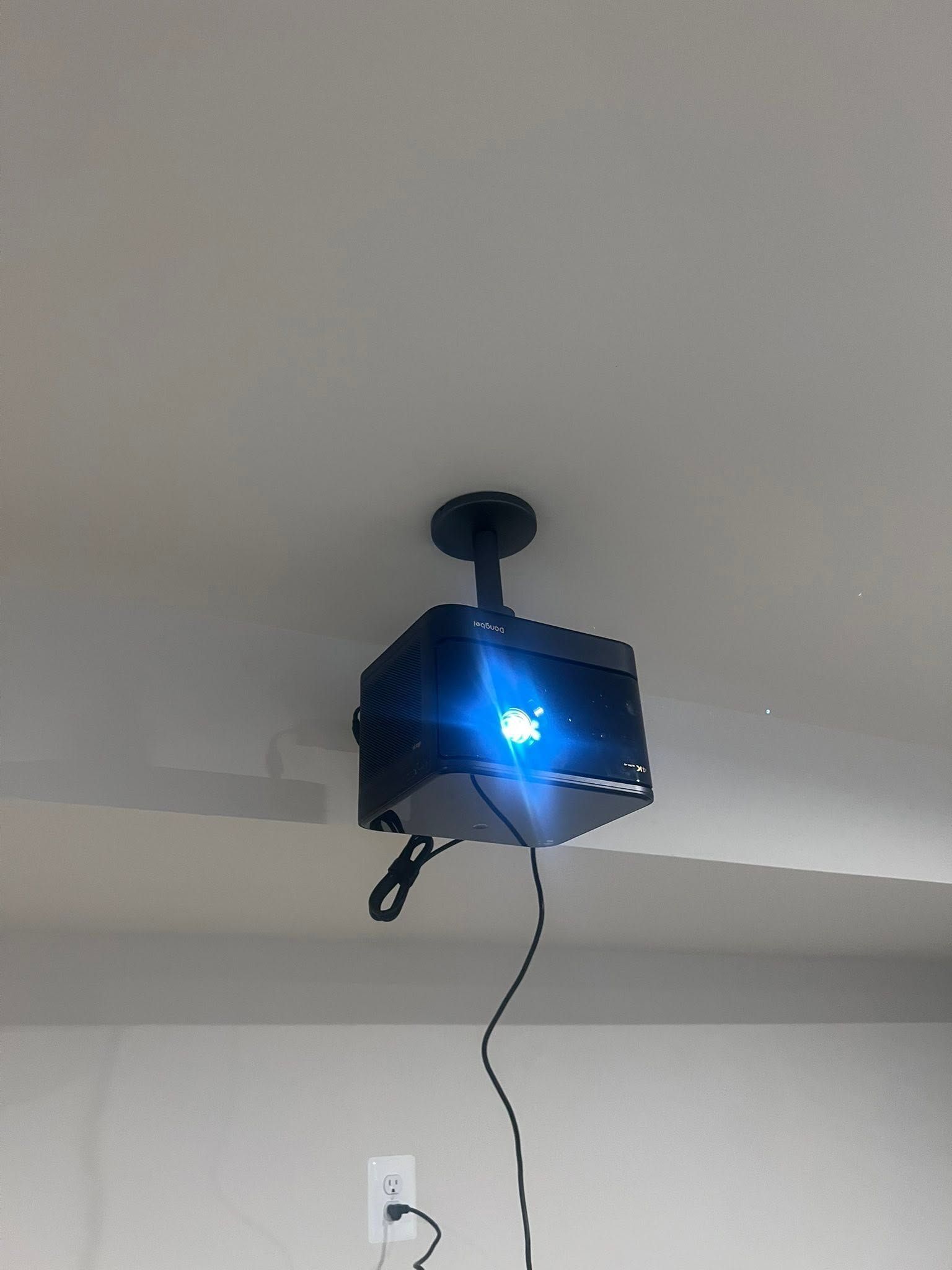 Black ceiling-mounted projector or light with a bright blue glow and a hanging cord on a white wall and ceiling.