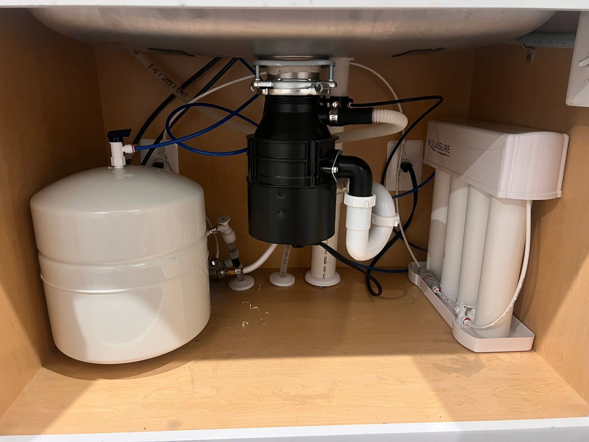 Under-sink plumbing with a white tank, black garbage disposal, and white water filter unit.