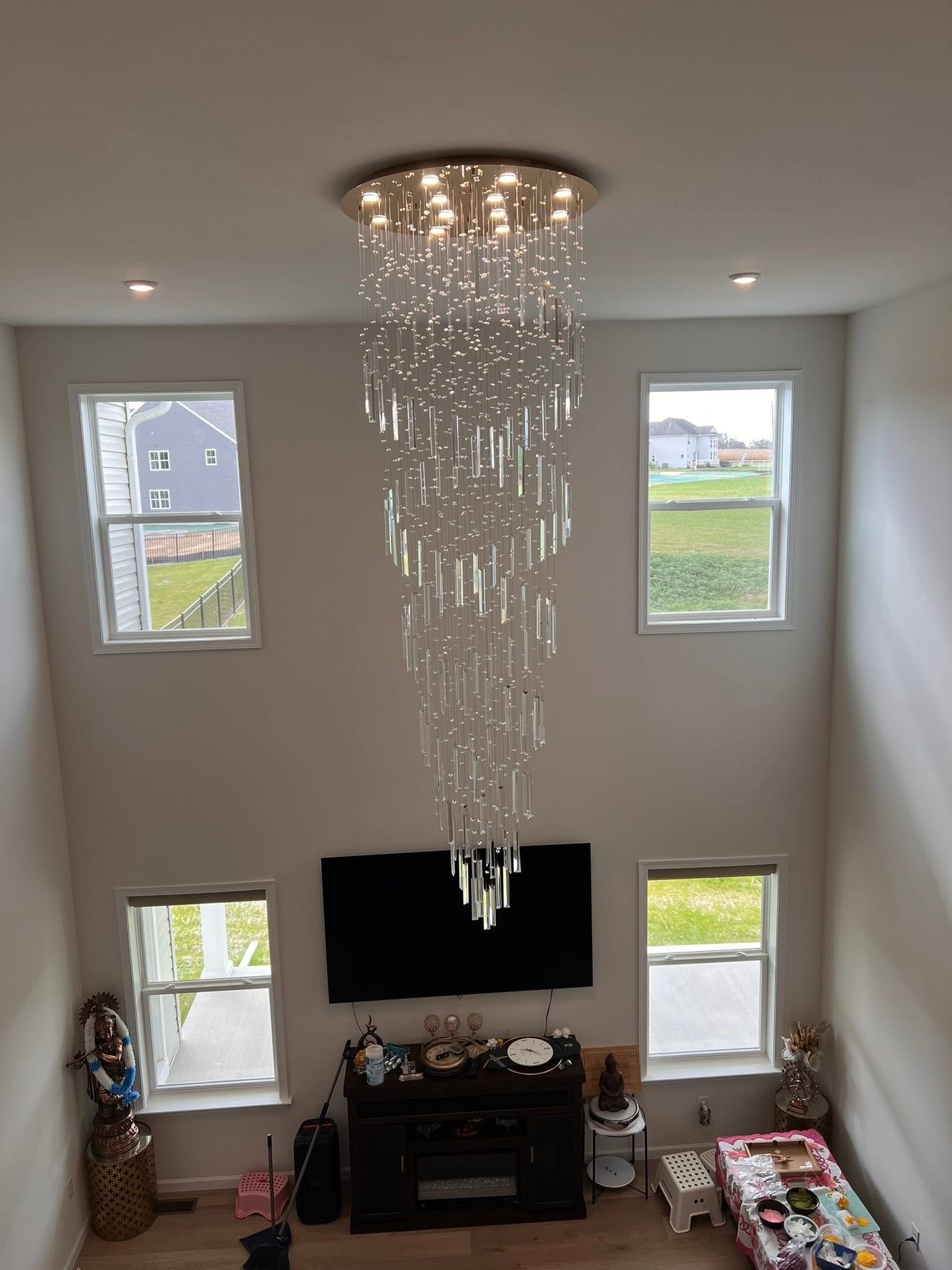 Tall chandelier hanging in a bright two-story living room with four windows and a TV below.