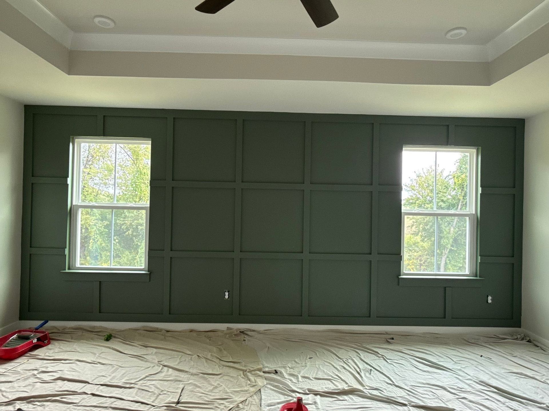 Green paneled wall with two windows in an unfinished room covered by protective floor paper