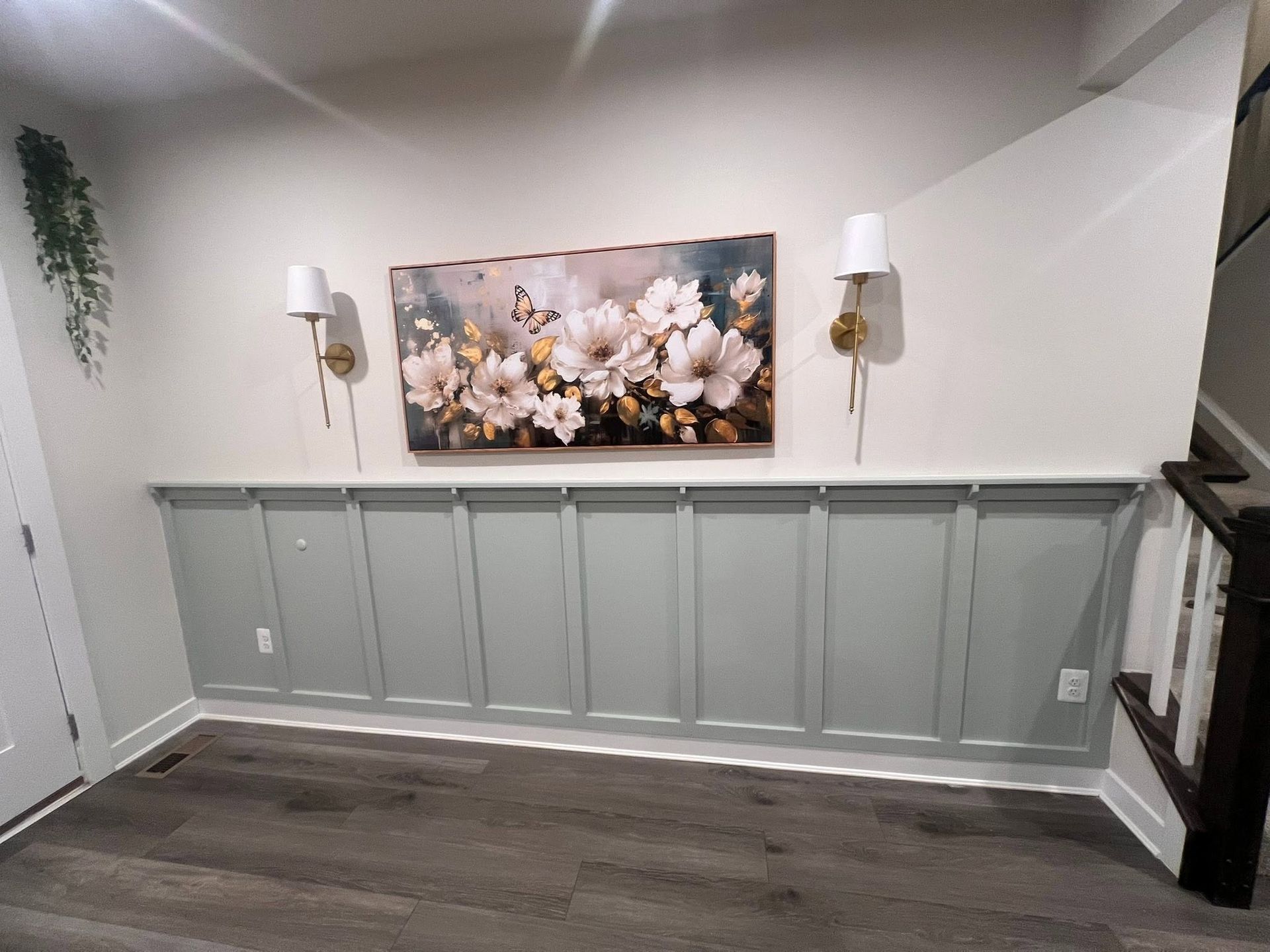 Gray hallway with floral wall art, white sconces, and paneled wainscoting above dark wood flooring