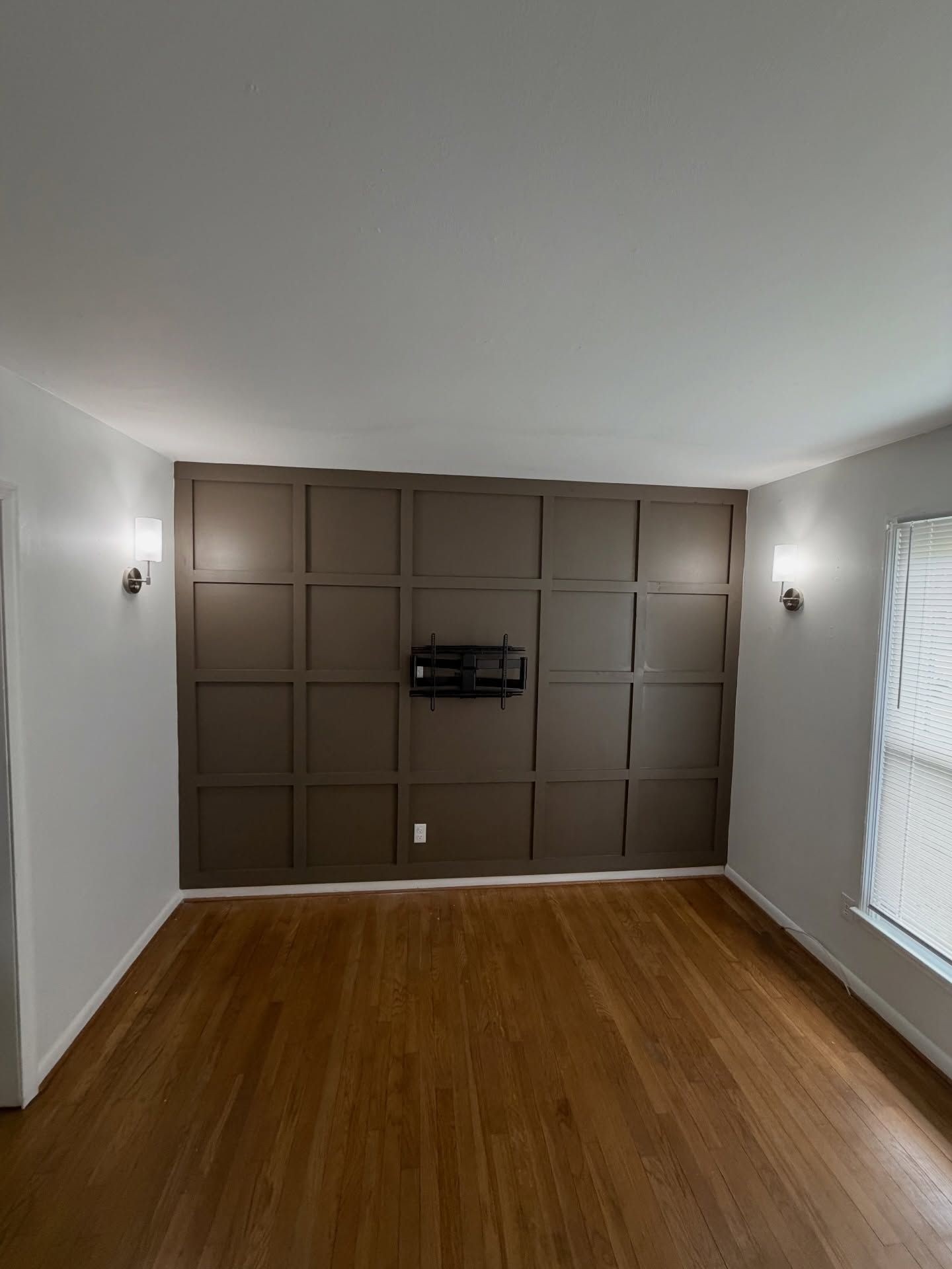 Empty living room with brown paneled accent wall, hardwood floor, and two wall sconces.