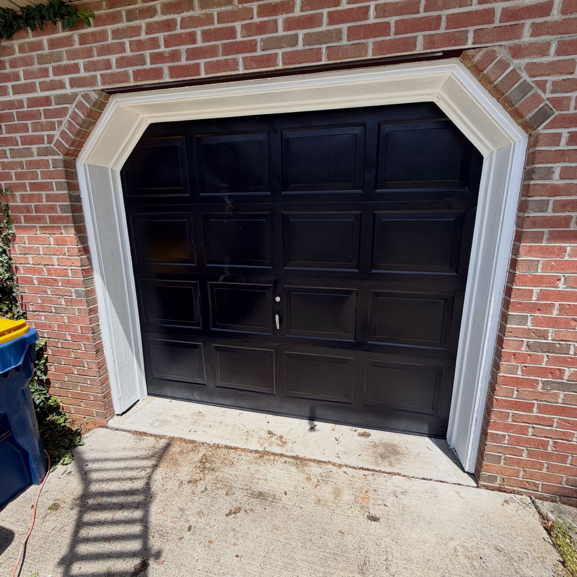 Black garage door in a brick house with a concrete driveway and a trash bin nearby