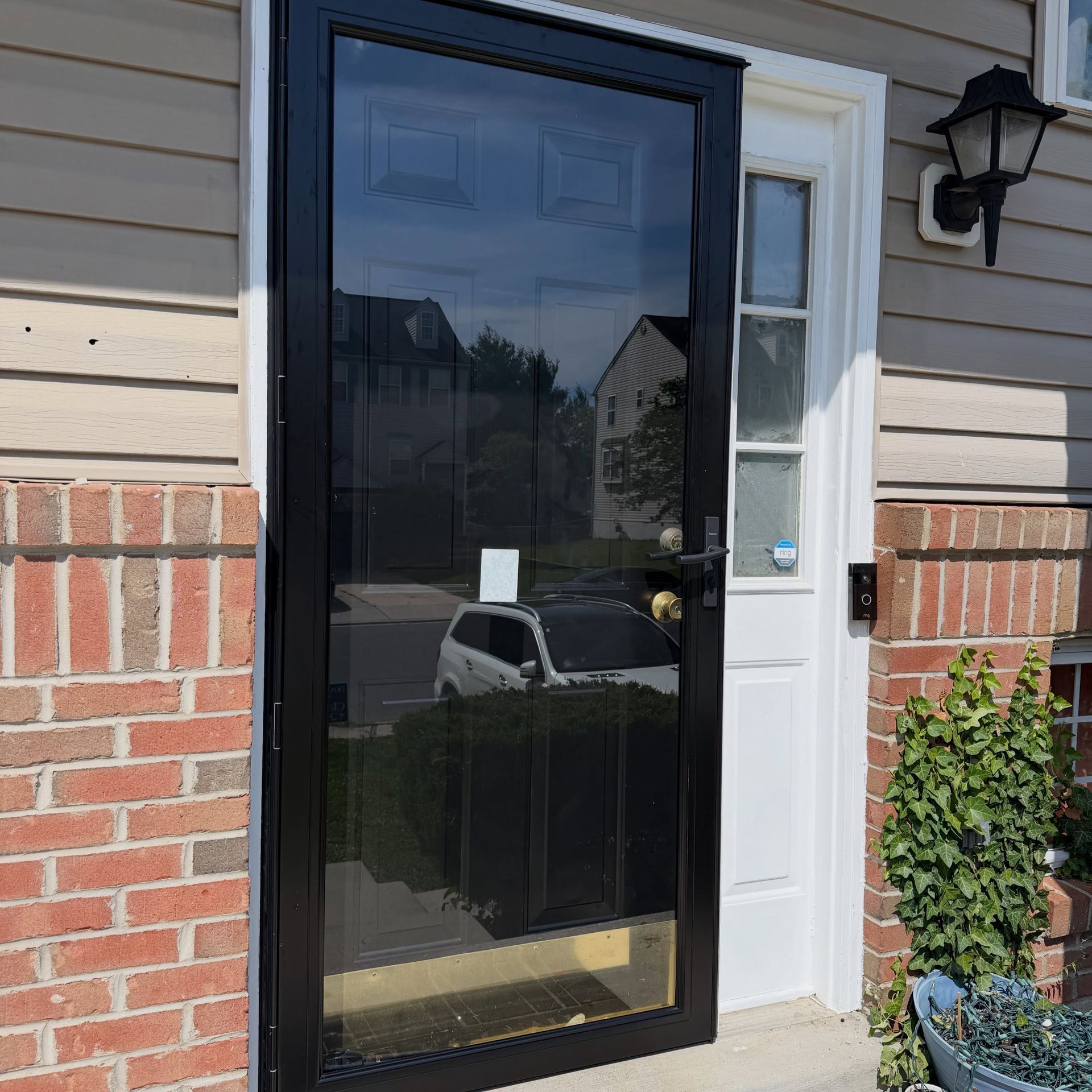 Black storm door beside a white entry door on a brick-and-siding house, with a wall lantern and potted plant.