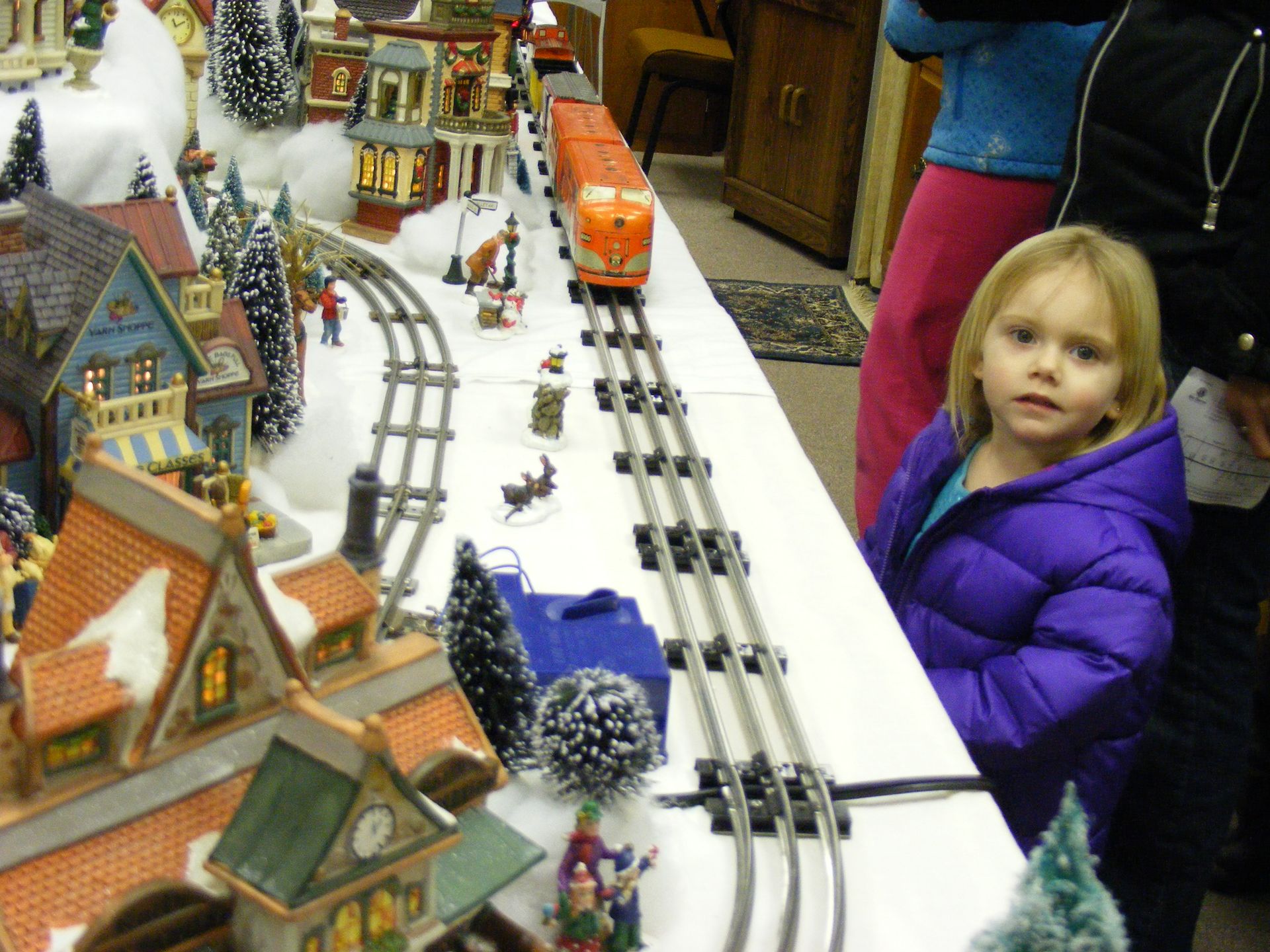 A young child watches a toy train travel through a miniature winter village scene.