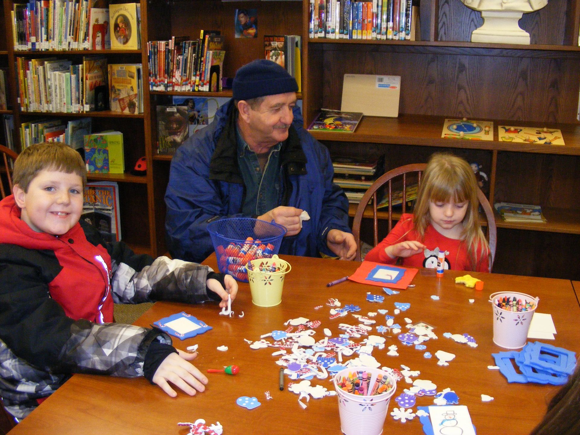 People at a table doing crafts in a library. A boy and girl are crafting with an older man. Smiling faces, colorful crafts.