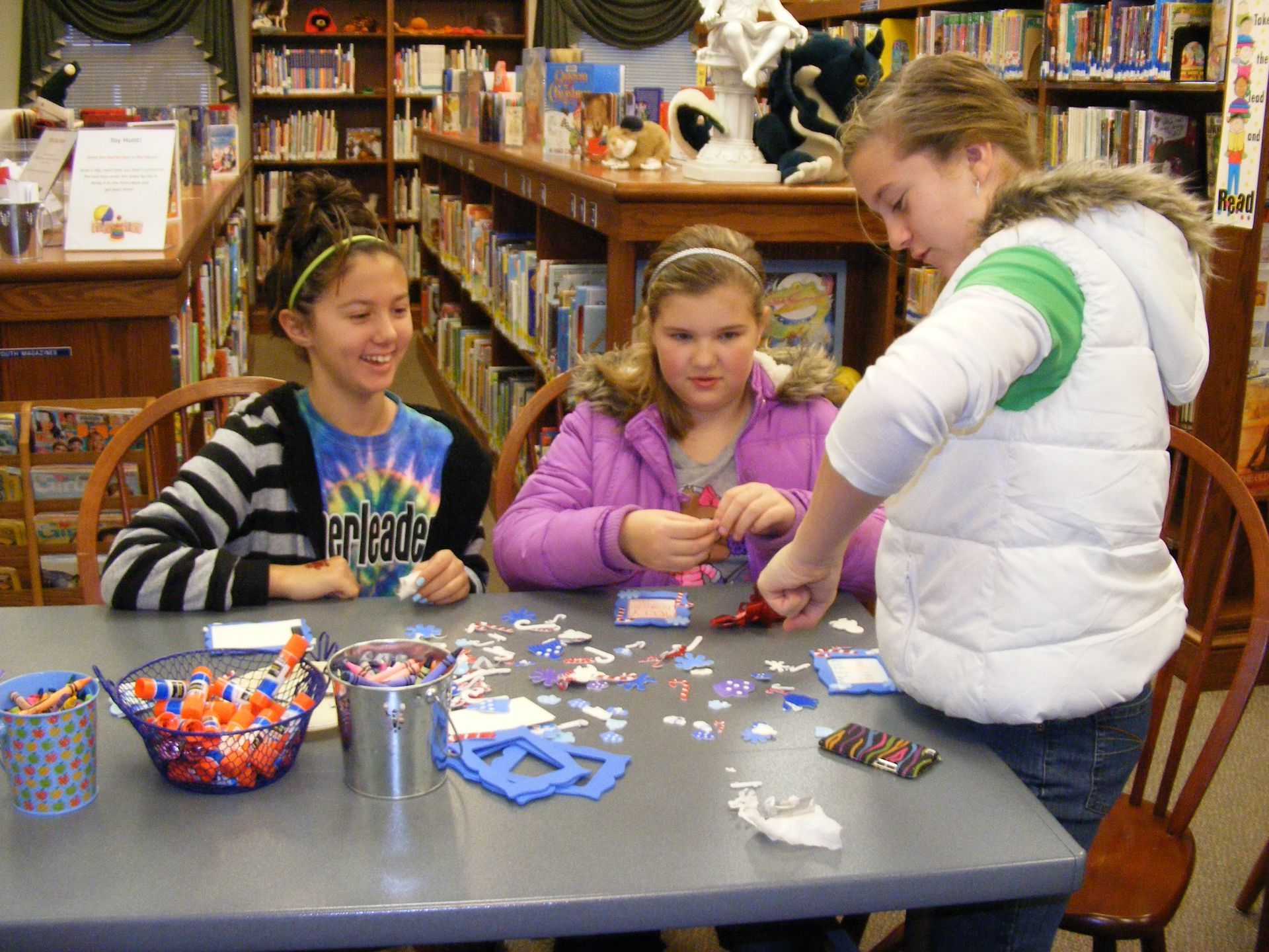 Three girls at a library table crafting with colorful materials.