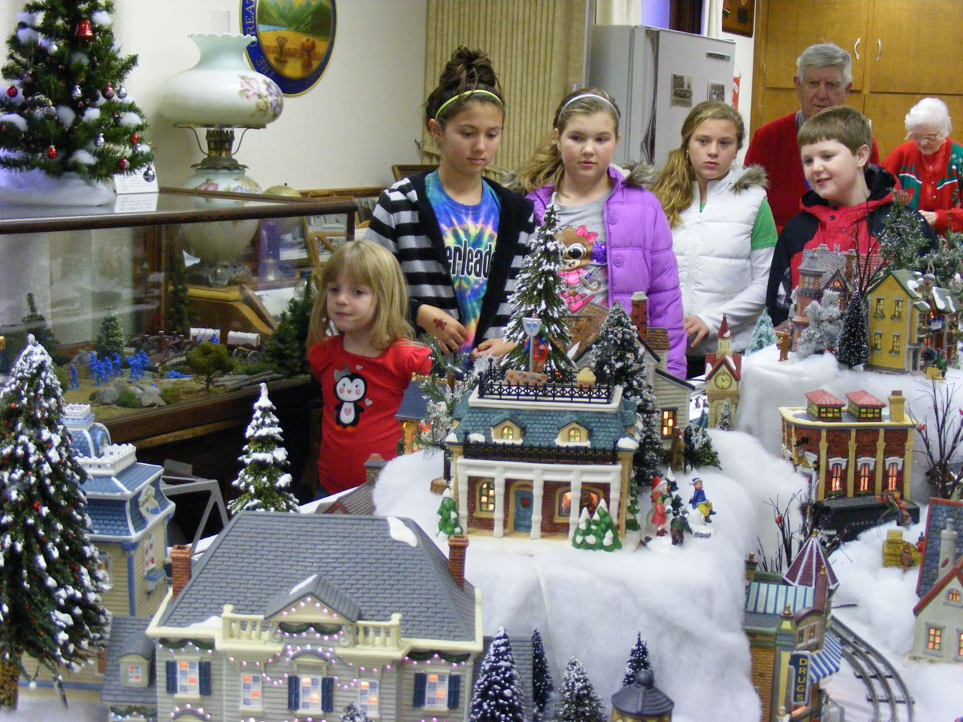Children looking at a snowy Christmas village display with miniature buildings and trees.