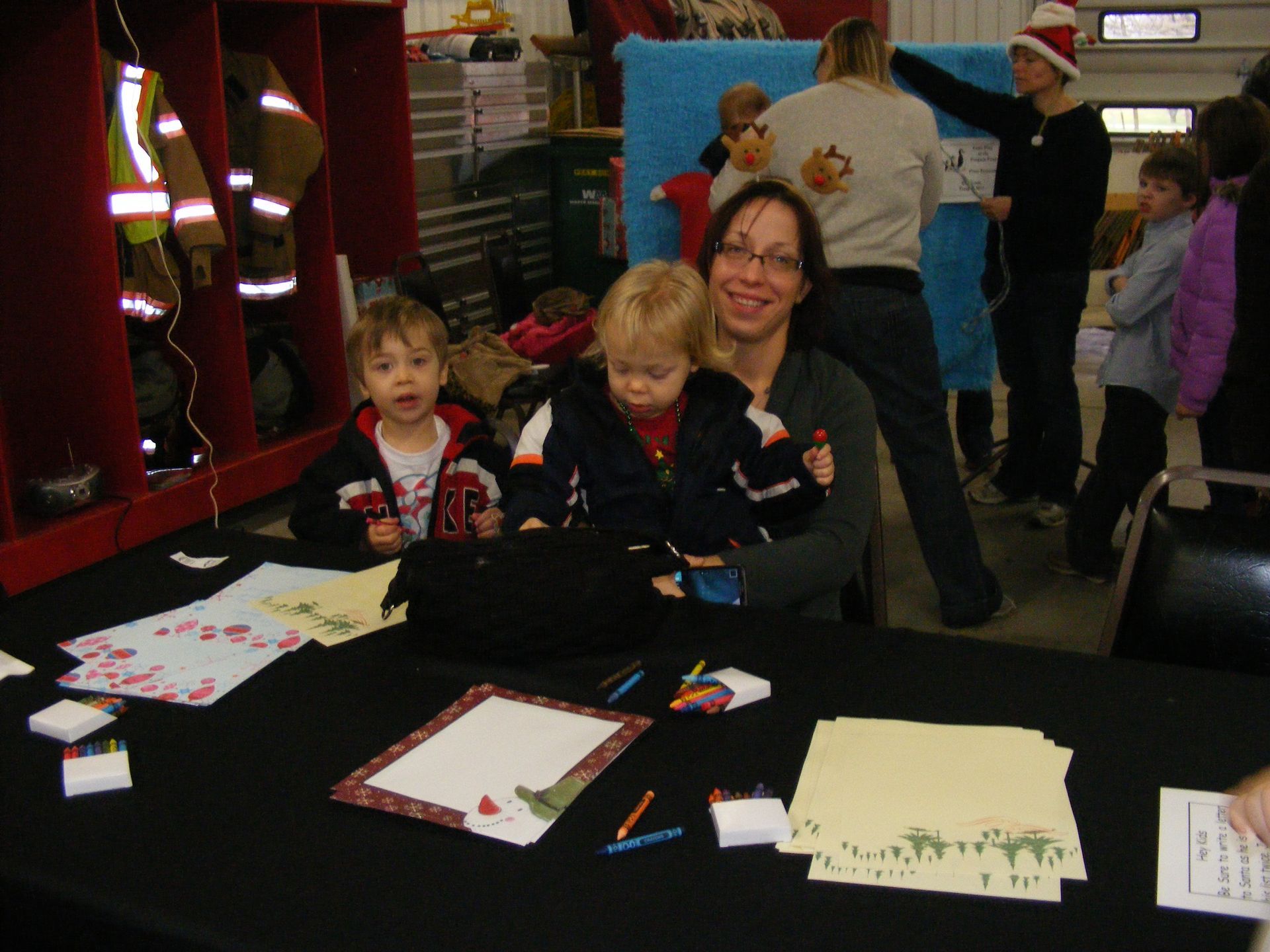 Woman and two young children at a craft table, smiling. Crayons, paper, and decorations are visible.