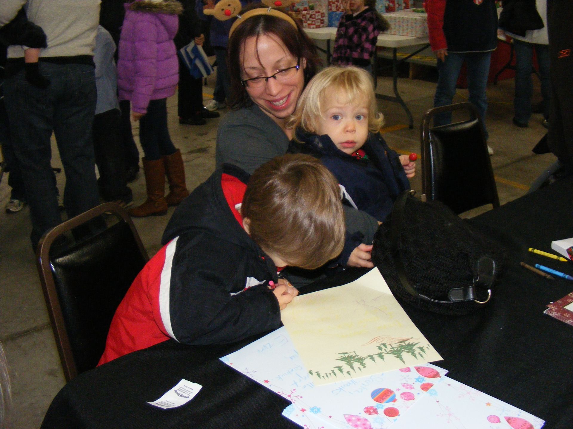 Woman seated, holding toddler, watching child draw at table; festive setting.