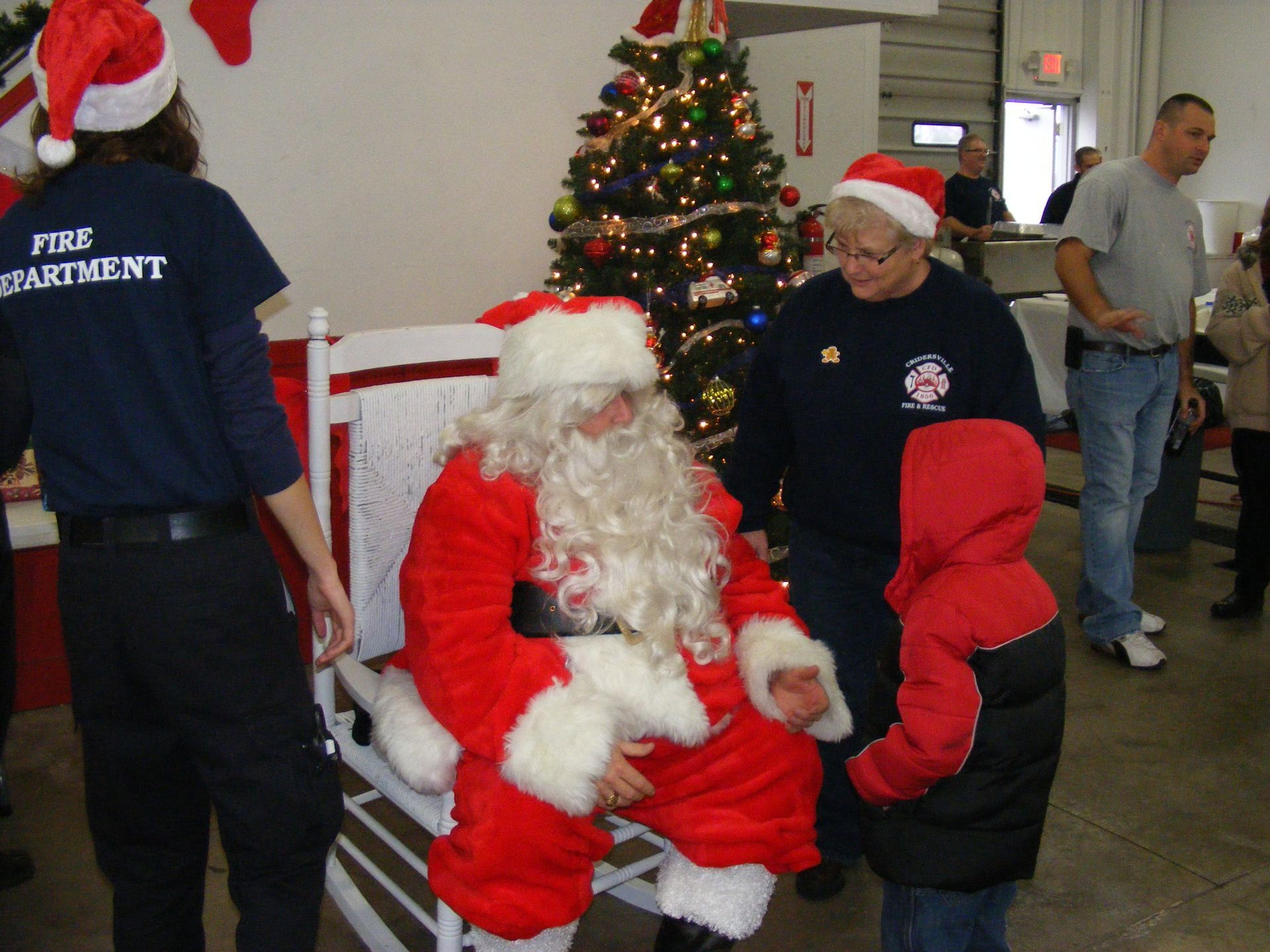 Santa Claus sits in a rocking chair with a child, surrounded by firefighters at a decorated Christmas tree.