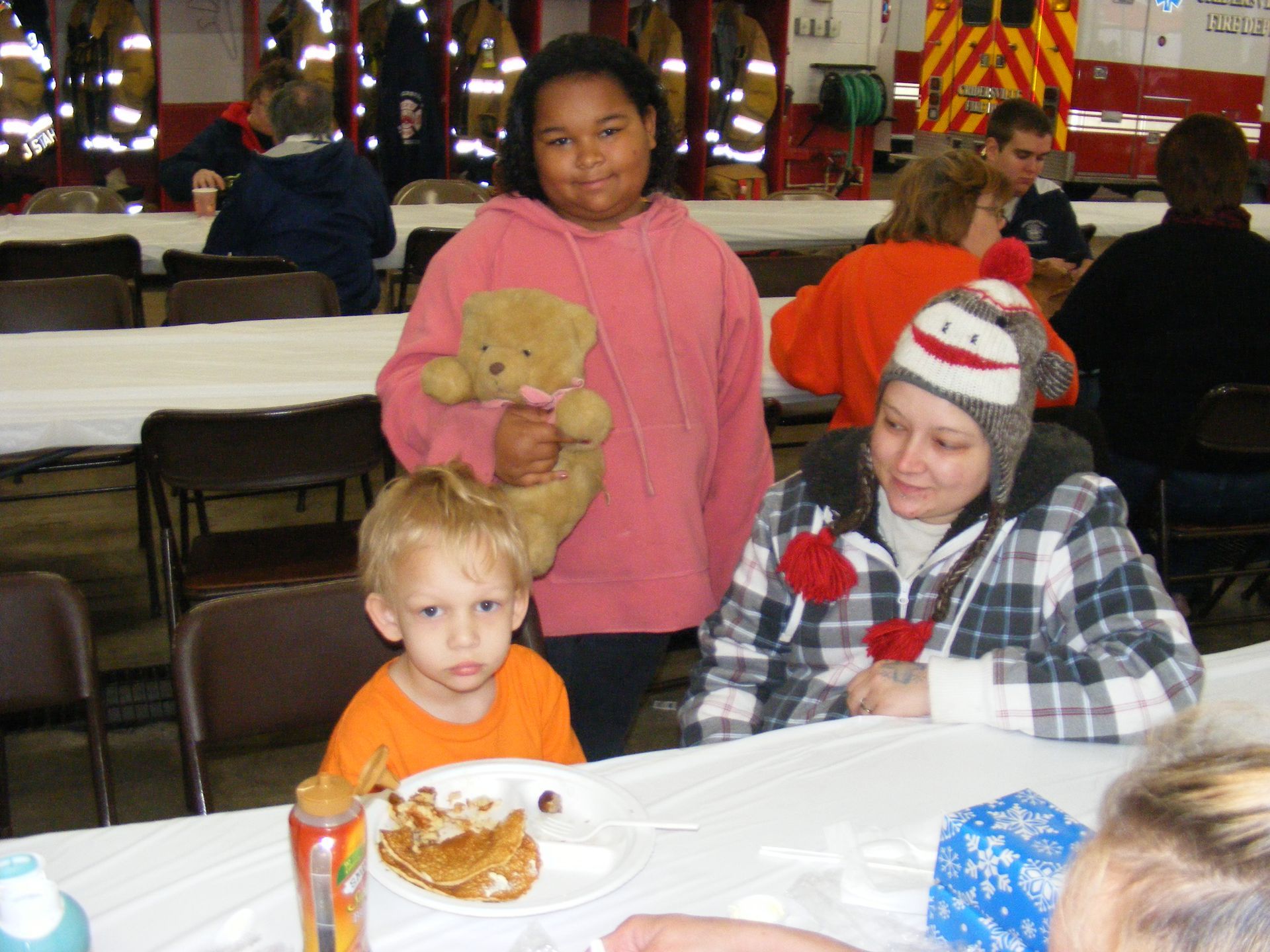 People at a table. A child with an orange shirt looks toward the camera, a girl holds a teddy bear, and a woman wears a hat.