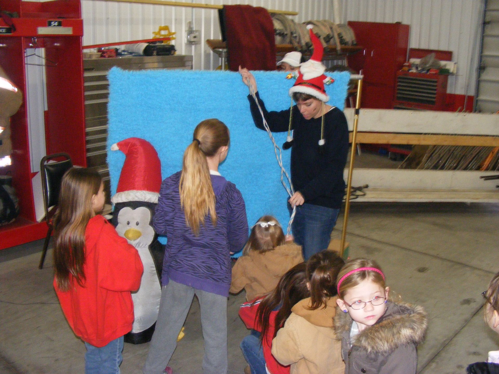 Children watch a person in a Santa hat, holding a stick in front of a blue backdrop, near an inflatable penguin.