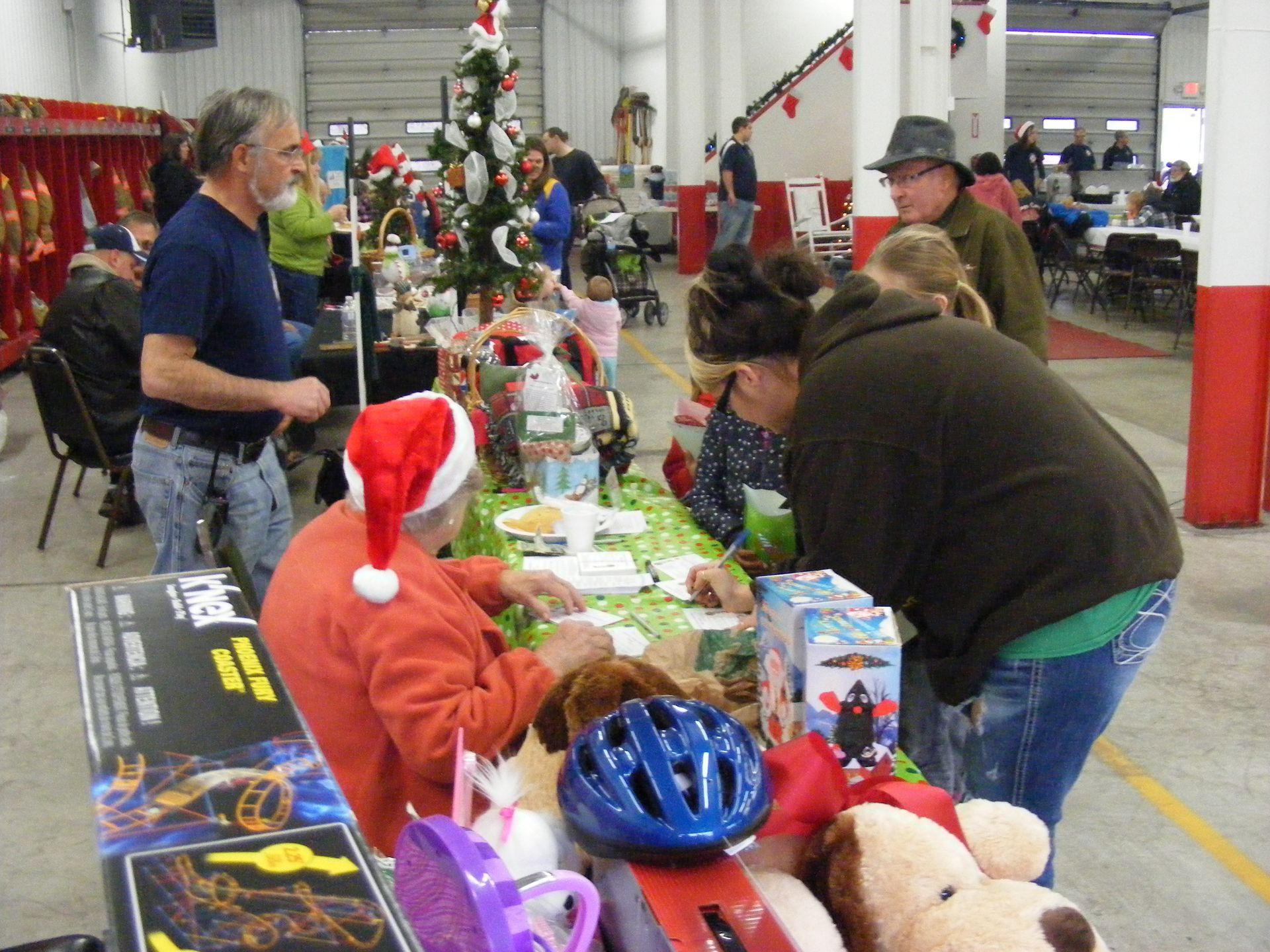 People browsing tables of gifts at an indoor holiday market, including a Christmas tree and toys.