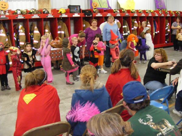 Children in costumes at a fire station, watching a performance. Red, pink, and orange costumes are visible.