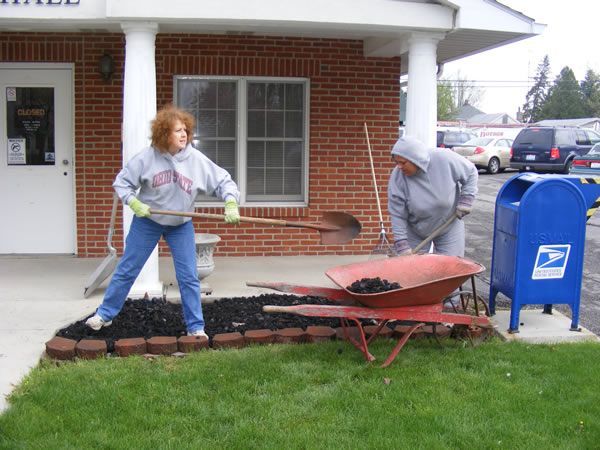 Two people mulching a flower bed in front of a building with a mailbox.
