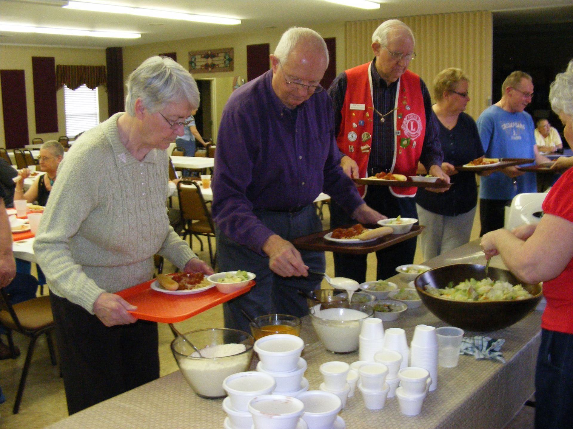 People serving themselves food from a buffet table at an event in a community hall.