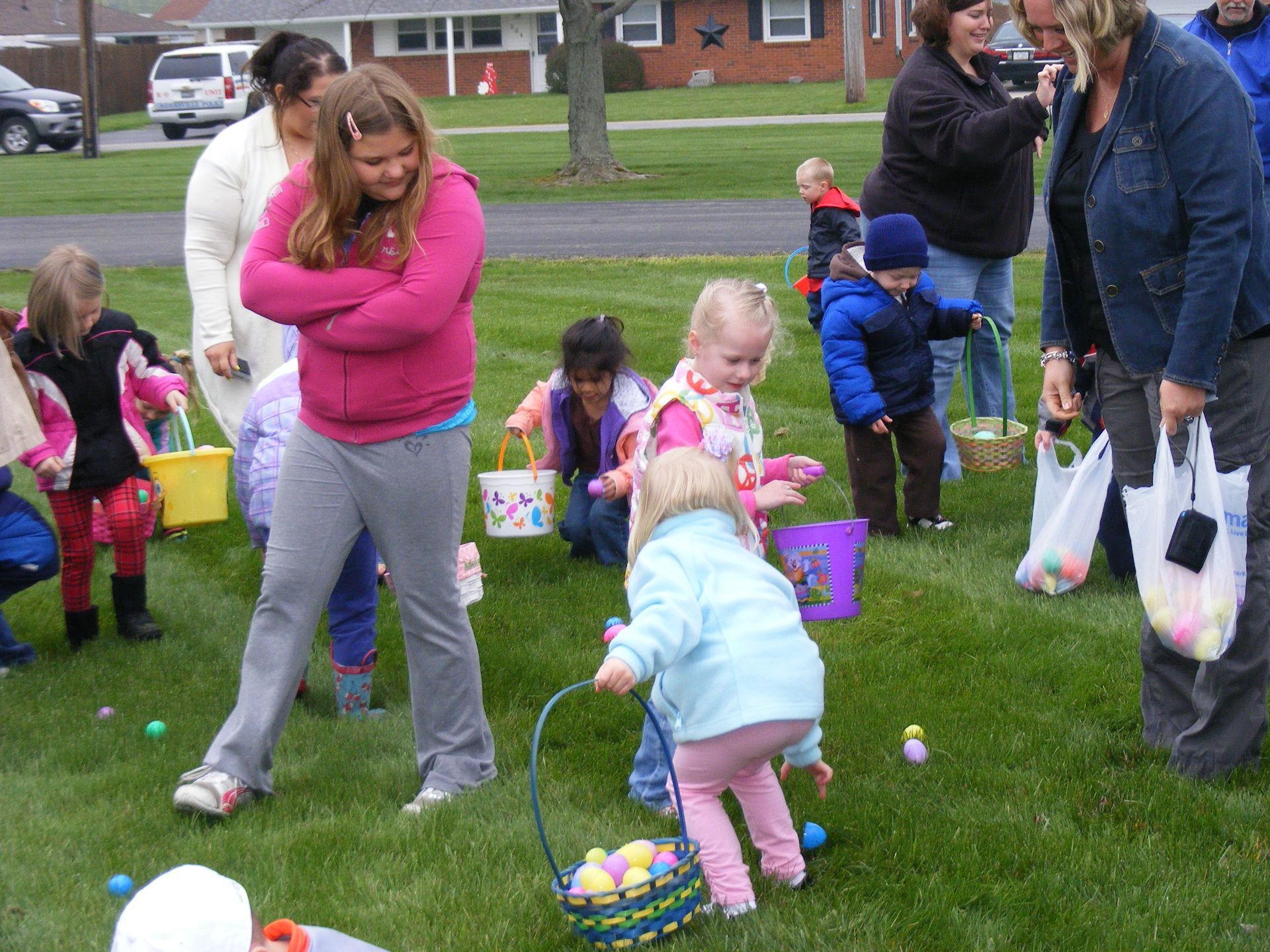 Children hunt for Easter eggs in a grassy field, surrounded by adults and baskets.
