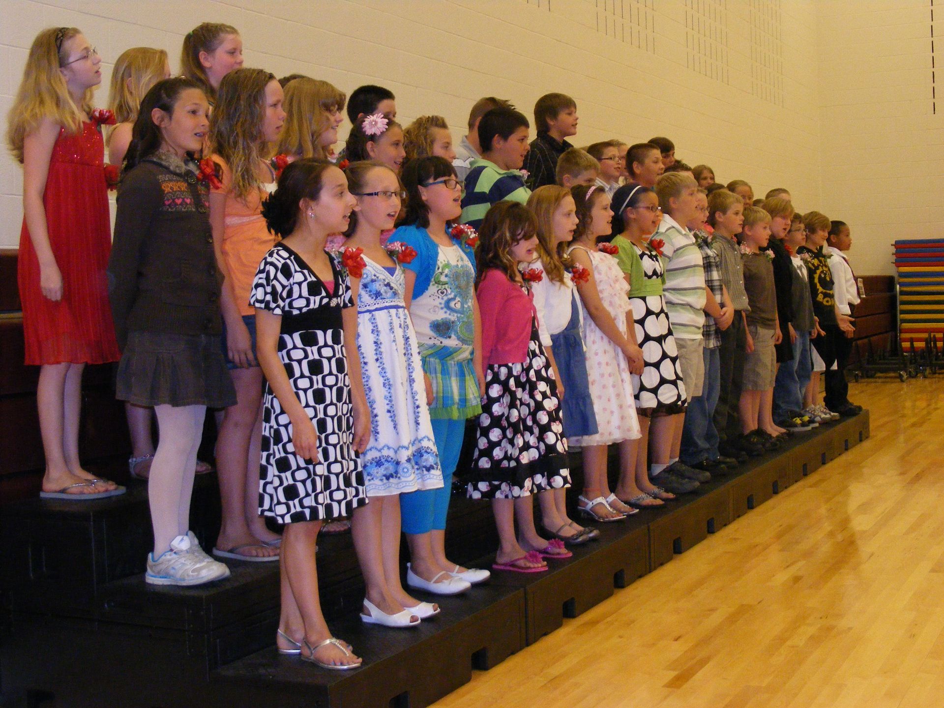 Children in colorful attire stand on tiered risers, possibly for a performance, indoors.