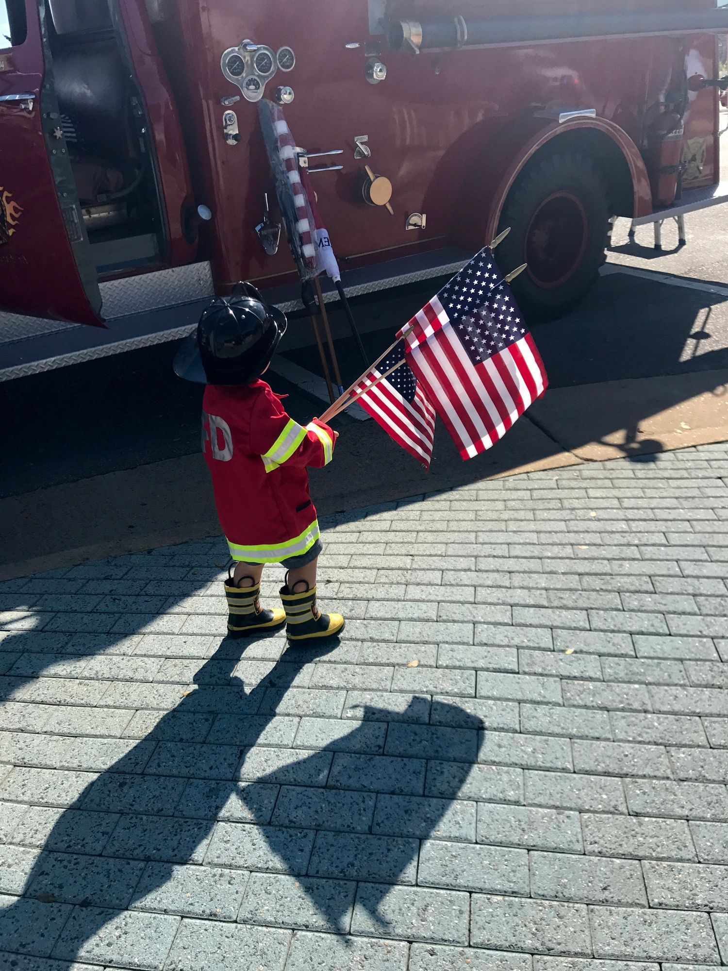 Child in firefighter uniform holds American flags near a red fire truck.