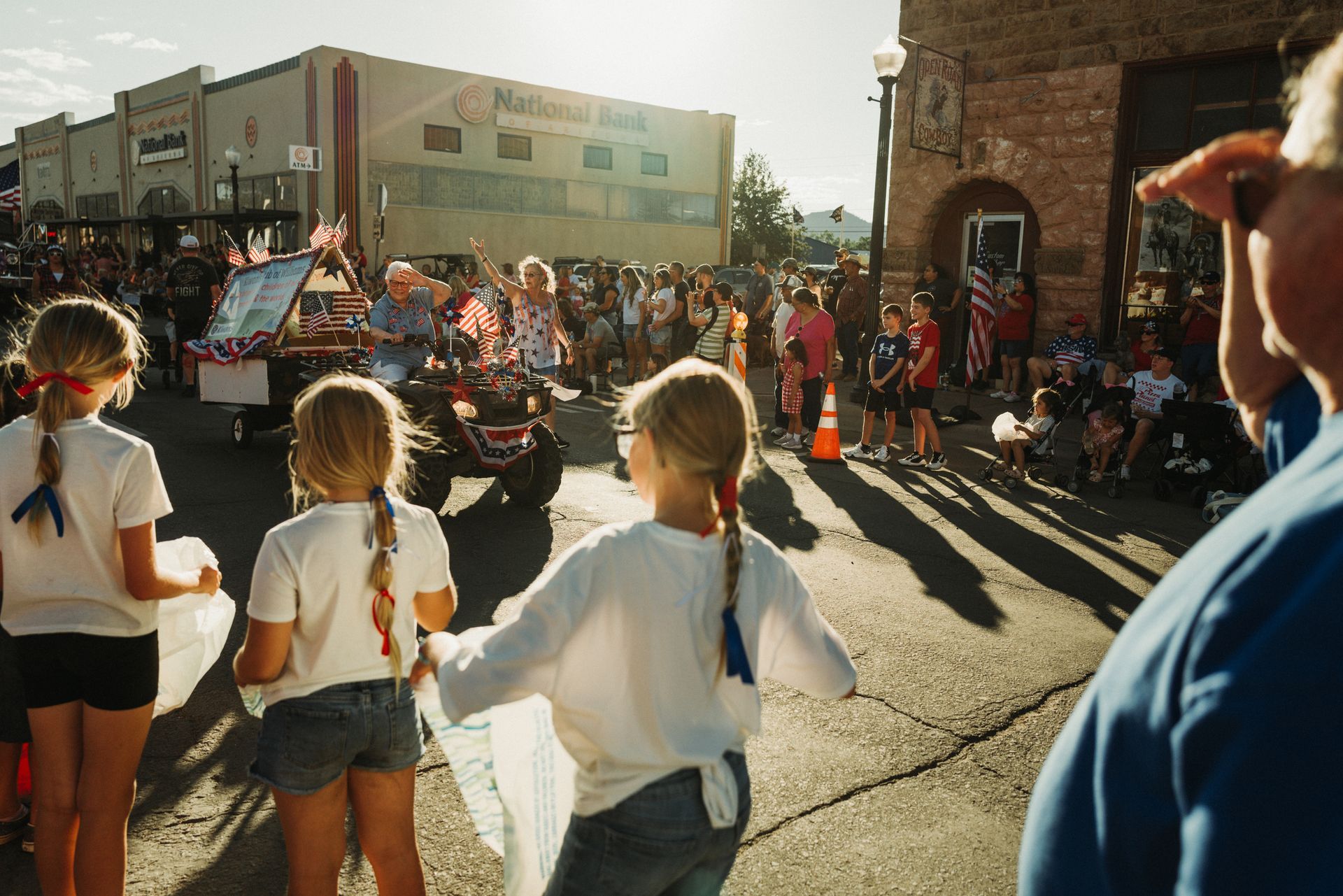 Children watch a parade on a street lined with buildings and people; golden sunlight.