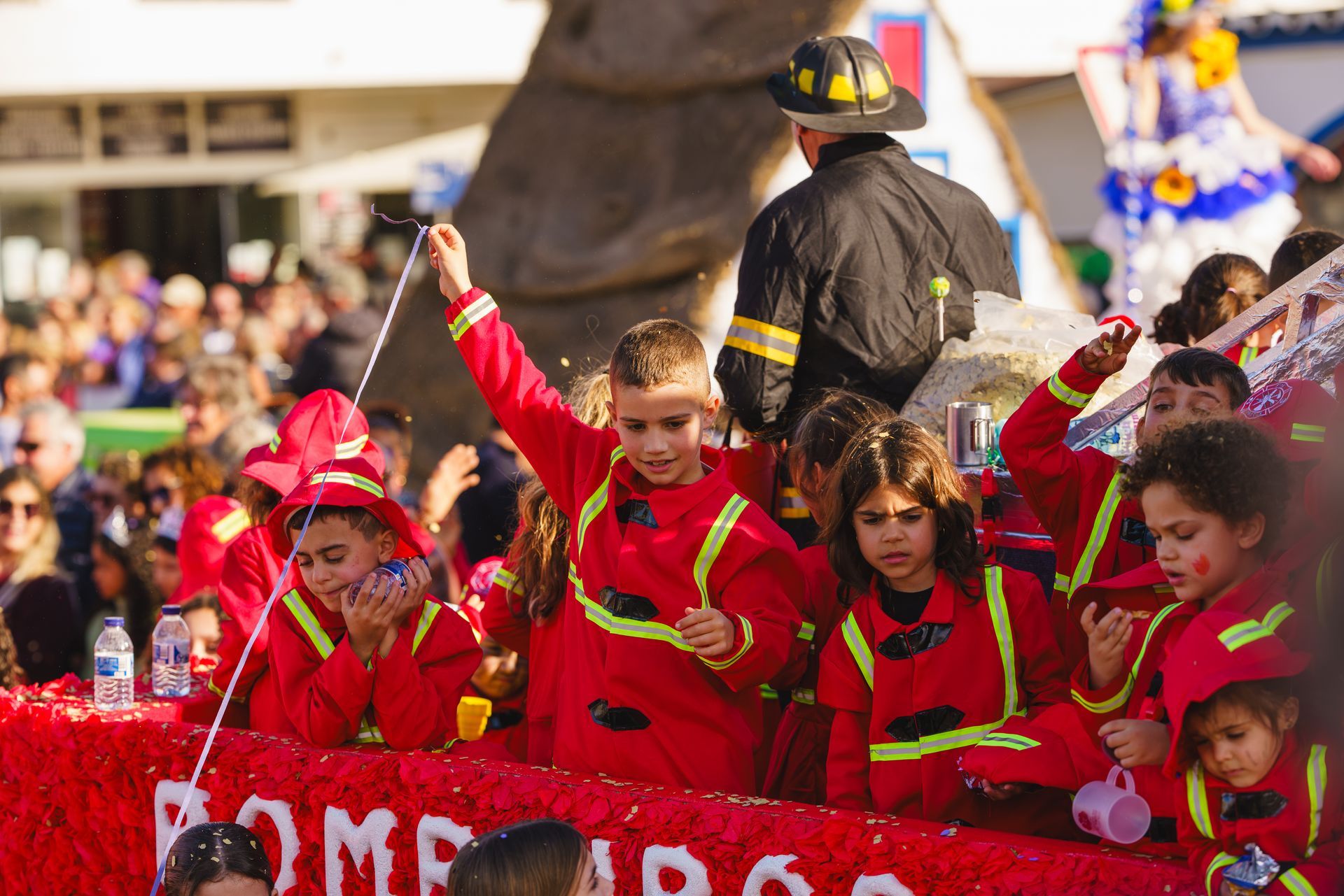 Children in red firefighter costumes on a parade float; a man in a firefighter uniform stands behind them.