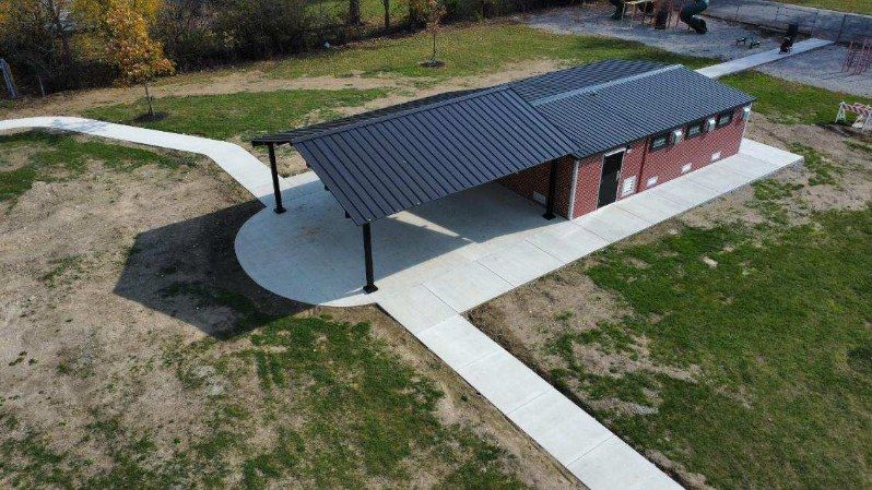 Park restroom with long, dark metal roof; concrete pathways; green grass.