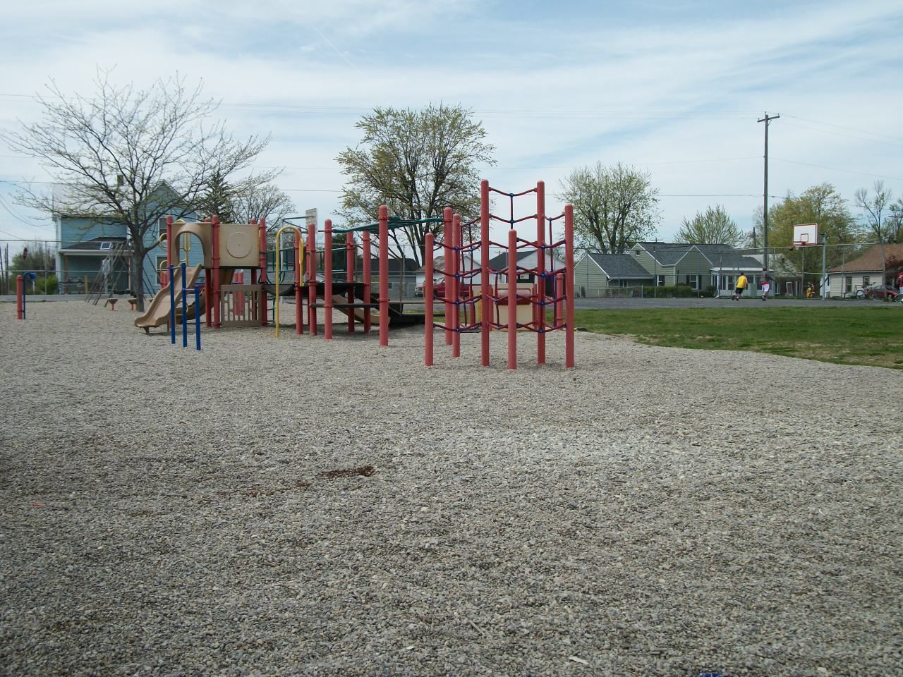 Playground with red and brown play structures on a wood chip surface, with trees and houses in the background.