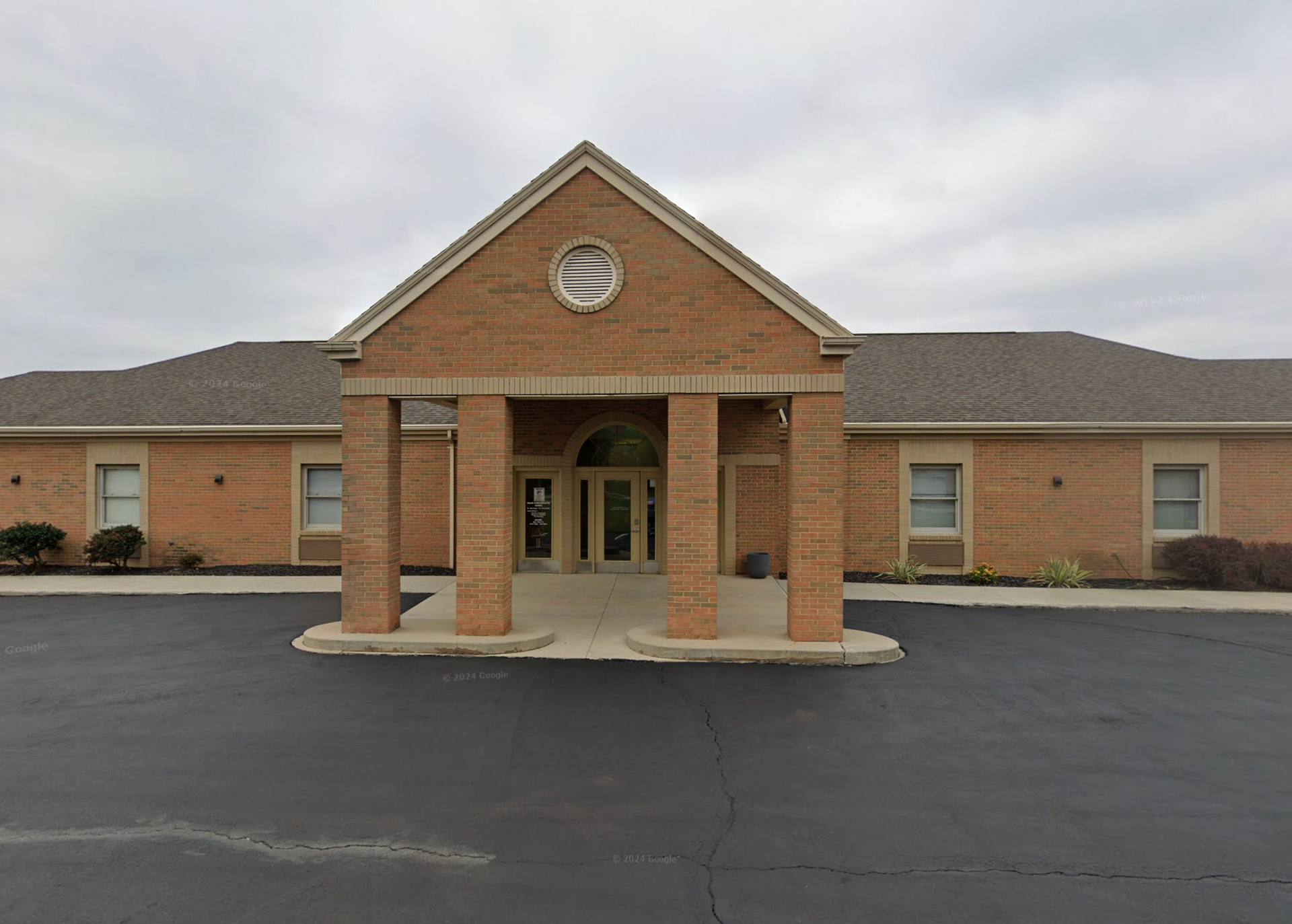 A brick building with a portico entrance and a dark paved parking lot in front under a cloudy sky.