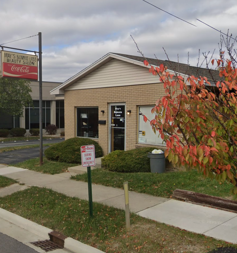 Exterior of a brick building with a barber shop sign, Coca-Cola sign, bushes, and a red-leafed tree.
