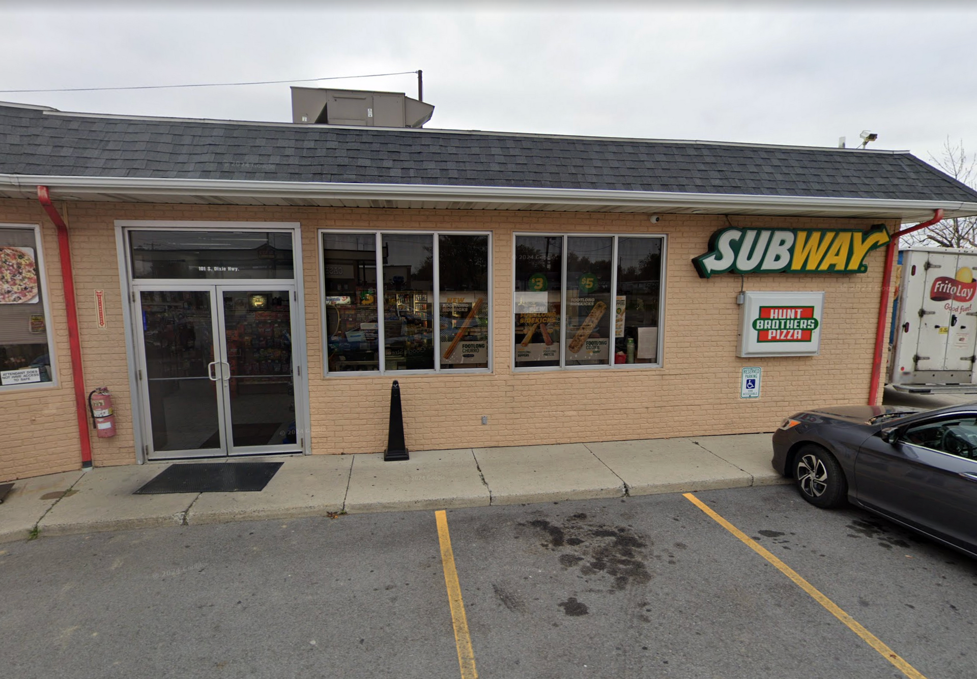 Subway restaurant with a black roof, beige brick facade, and a parked gray car.