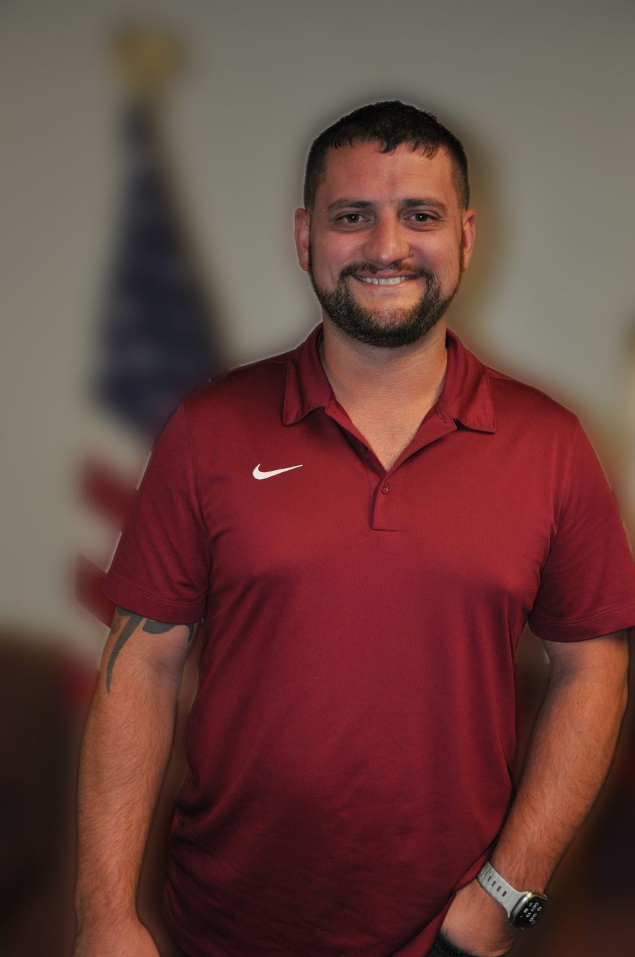 Man in red polo shirt smiles, blurred American flag in background.