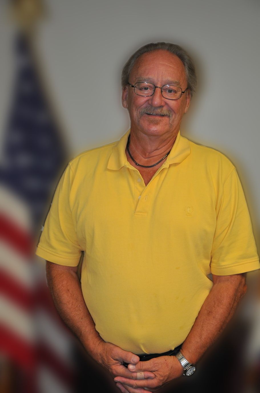 Man in yellow polo shirt, standing, with hands clasped; American flag in the background.
