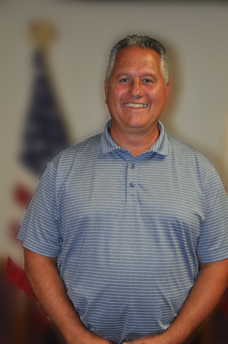 Man in blue striped polo smiling, standing indoors near a blurred flag.