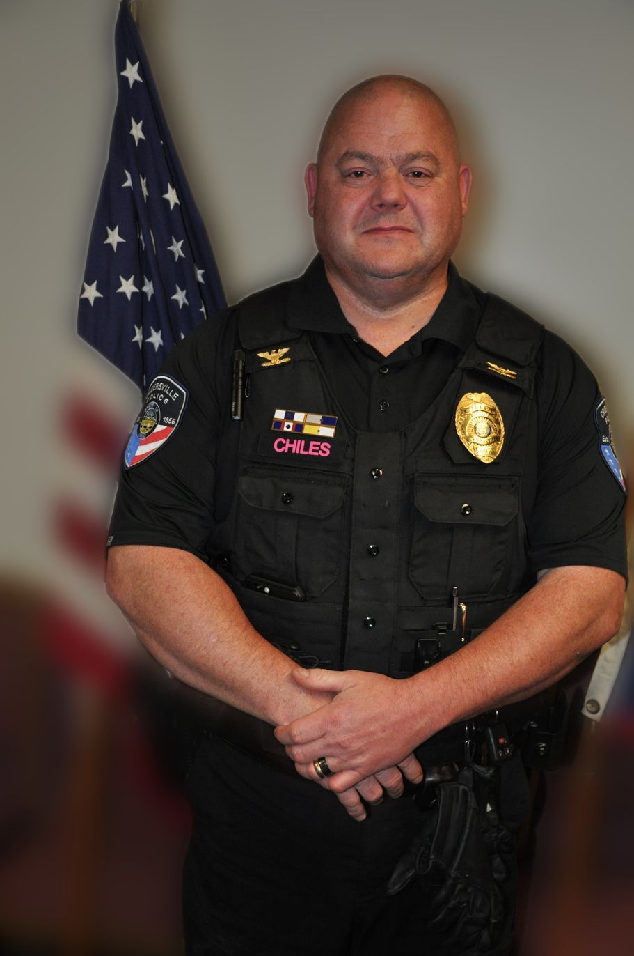 Police officer in black uniform, standing in front of American flag, arms crossed.