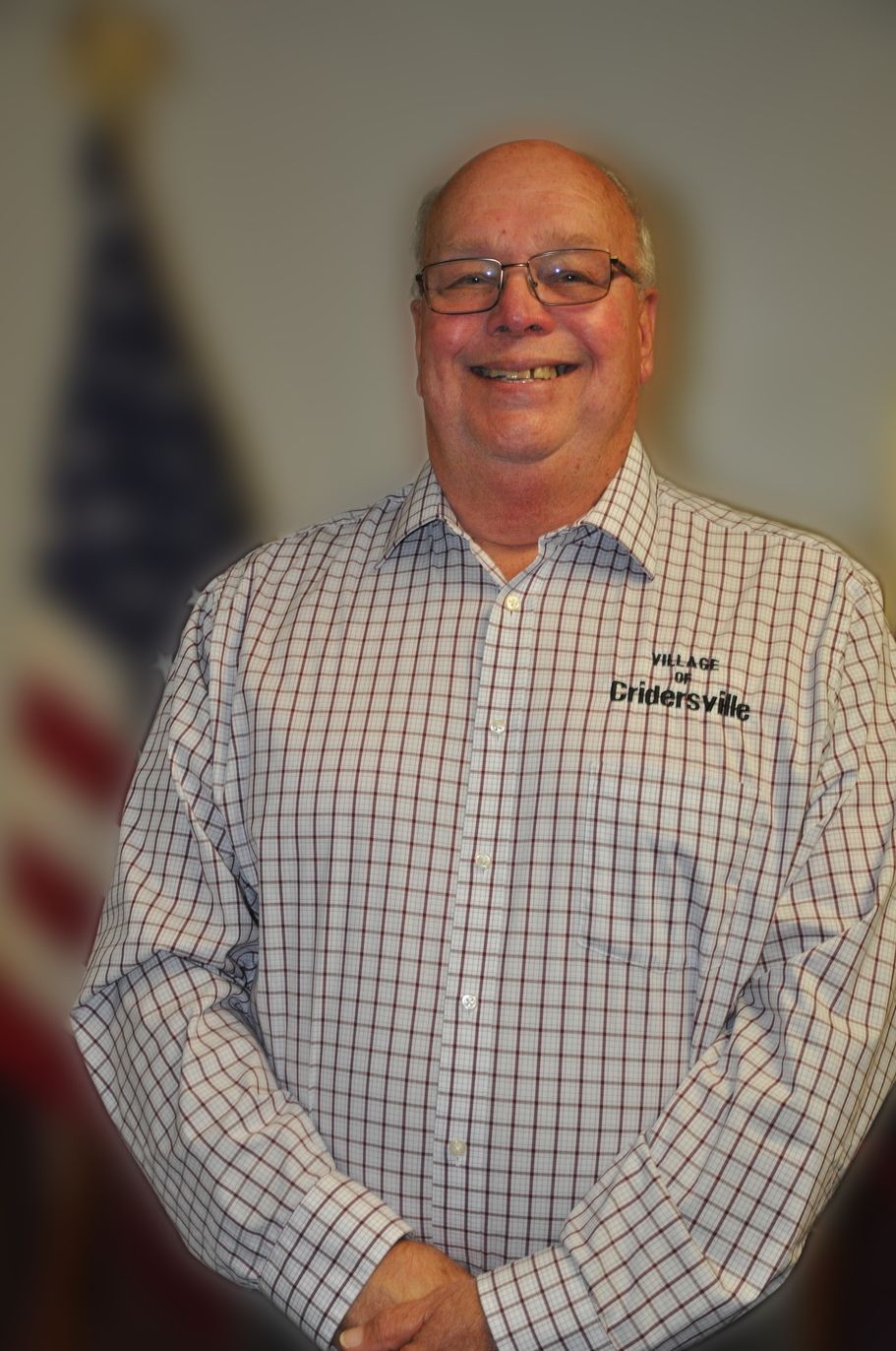 Man smiling, wearing glasses and a checkered shirt. The American flag is blurred in the background.
