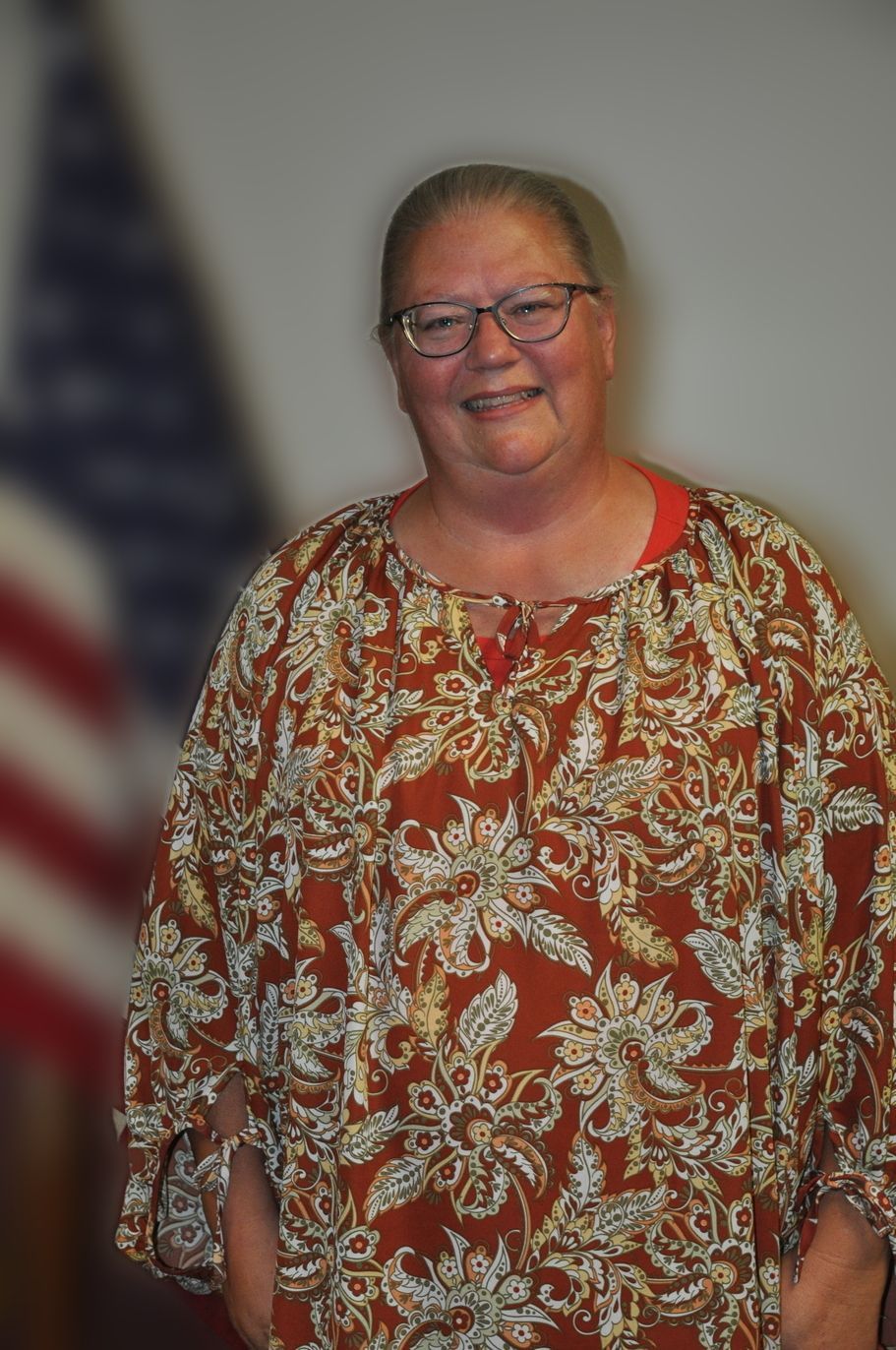 Woman wearing glasses and a patterned top smiles in front of a blurred American flag.
