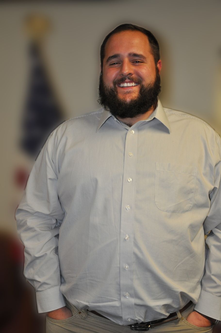 Man with a full beard smiling, wearing a button-up shirt, hands in pockets. Blurred background.