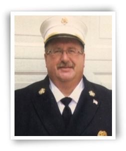 Man in fire department uniform, wearing a white hat, black jacket, and tie.