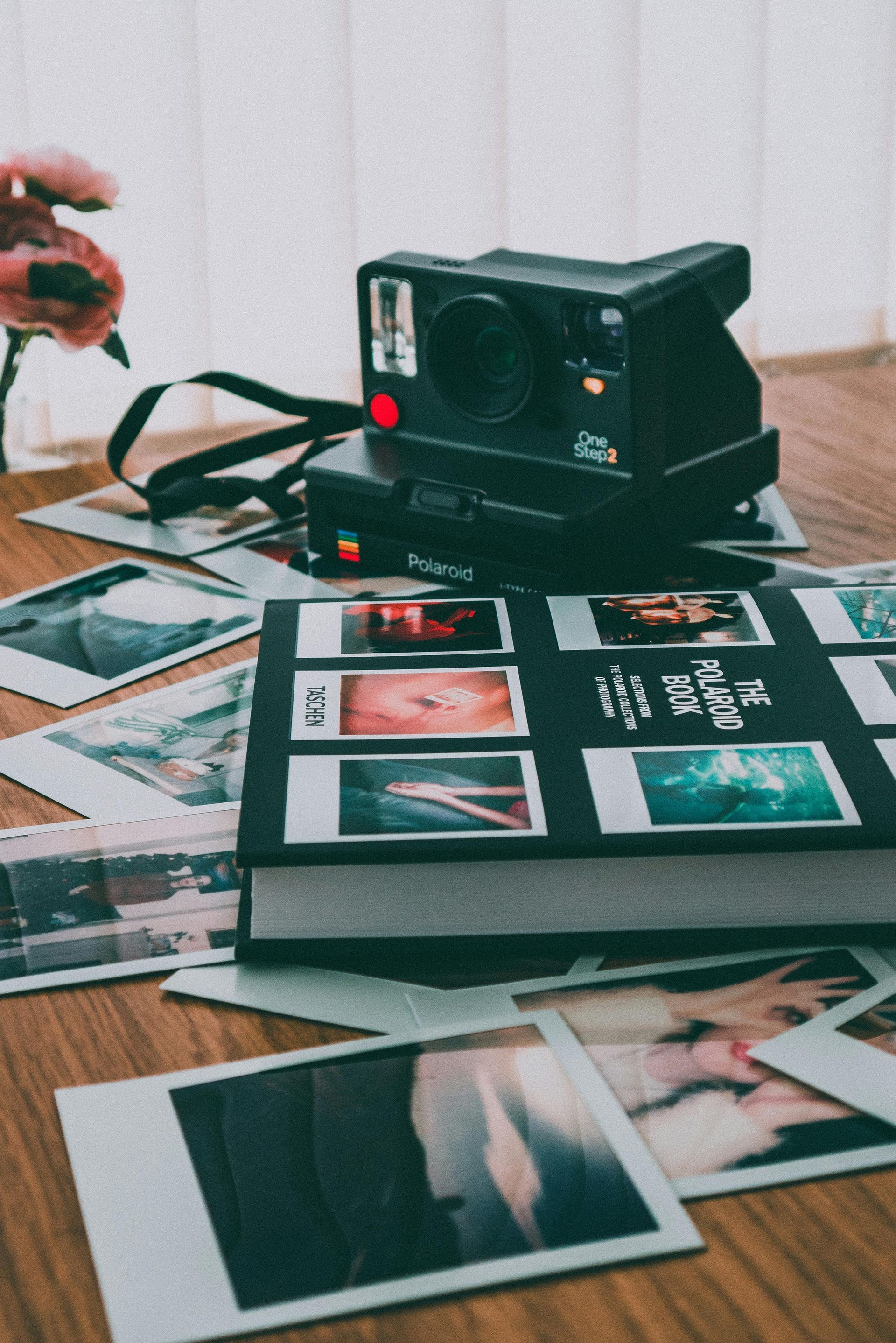 Polaroid camera on a table with scattered photos and a book; flowers in the background.